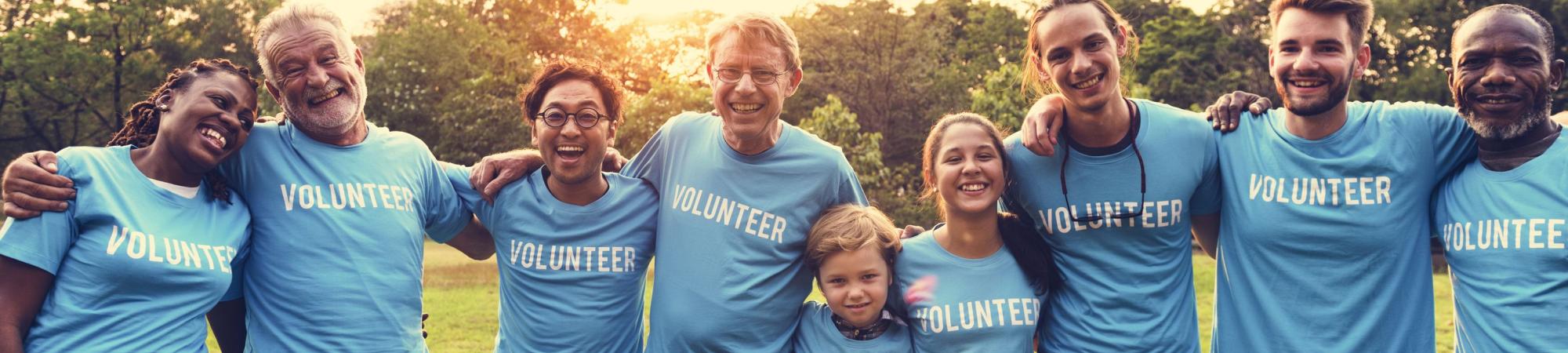 A group of volunteers of various ages stand in a line with their arms around each other's shoulders and smile at the camera